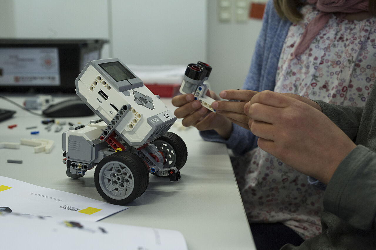 Zwei Personen sitzen am Tisch und bauen an einem Lego-Roboter mit Rädern. Es sind nur die Hände und der Roboter zu sehen.